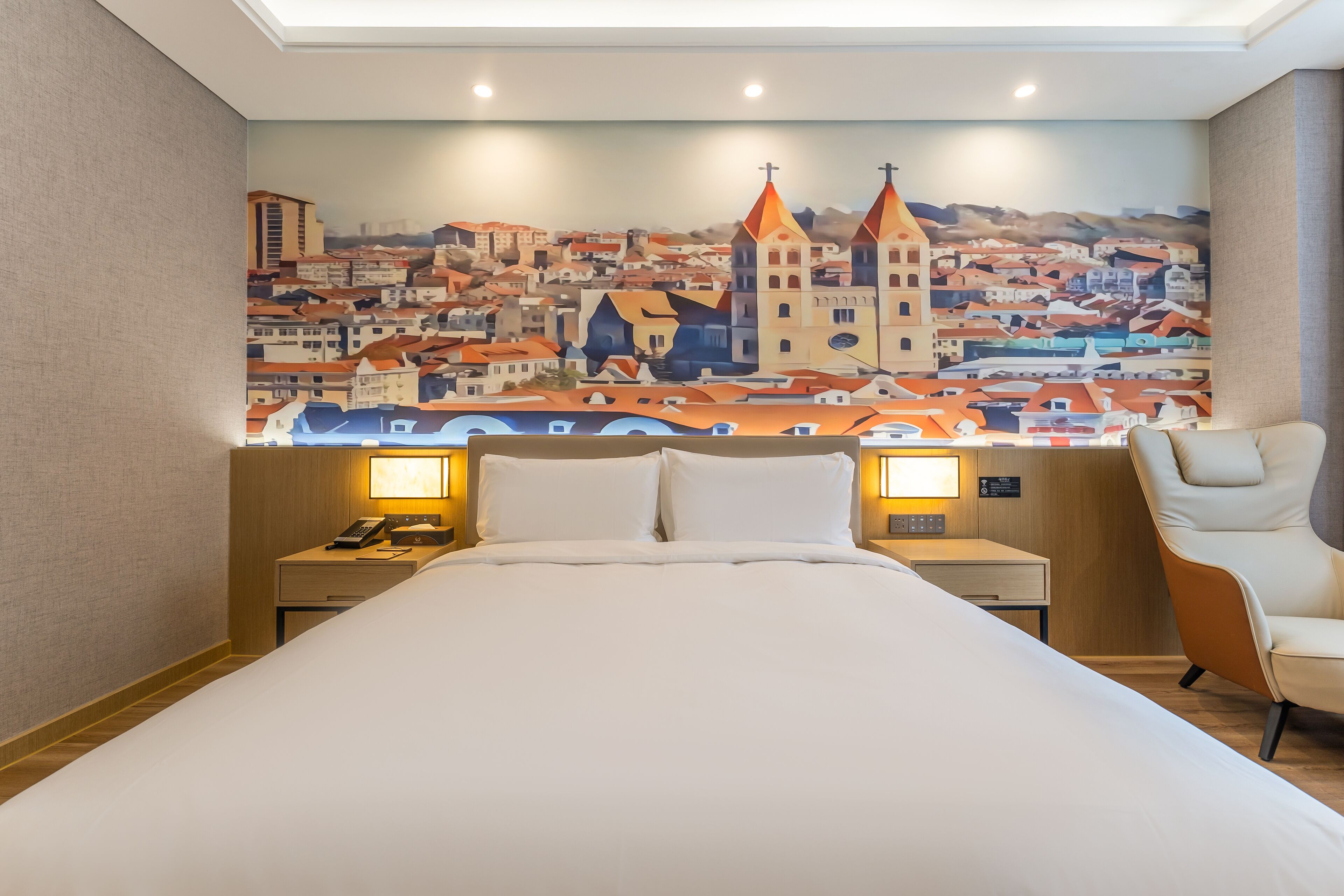 Habitación de hotel con una cama grande, mesitas de noche con lámparas, una silla y un mural con una vista urbana en la pared.