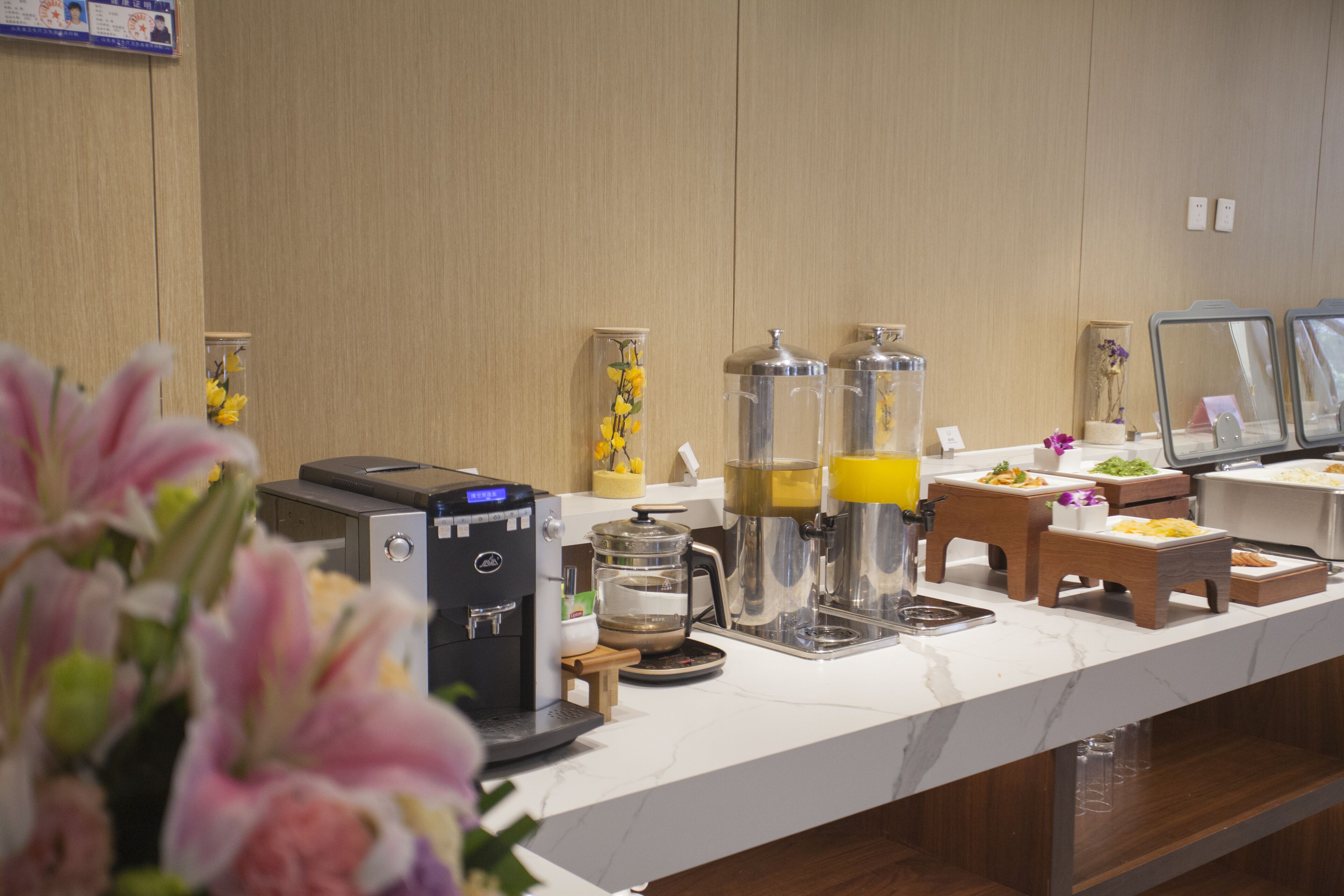 Zona de recepción de un hotel con una máquina de café, dispensador de agua y un pequeño rincón de estar con un arreglo floral.
