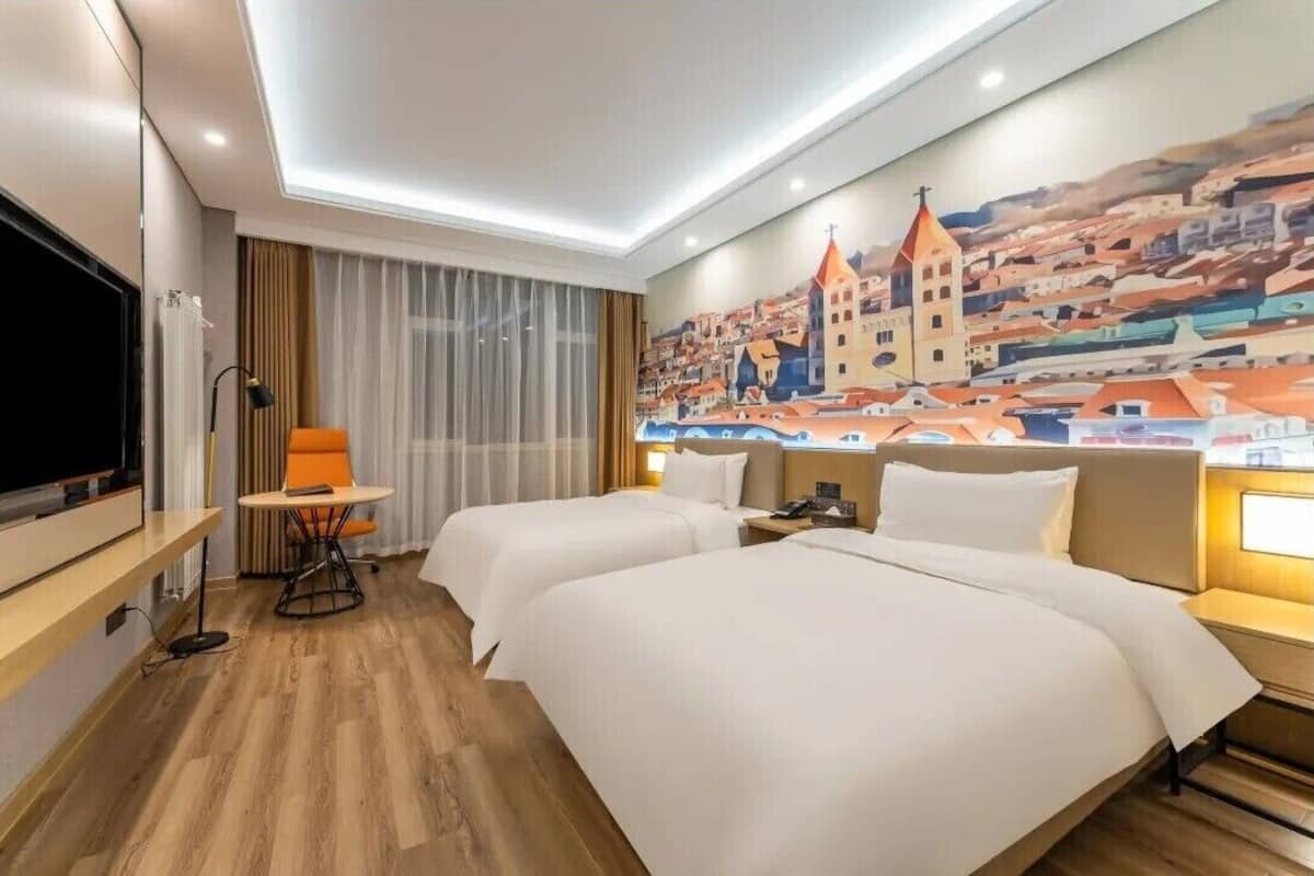 A hotel room with two beds, a TV, a desk, and a painting of a town.