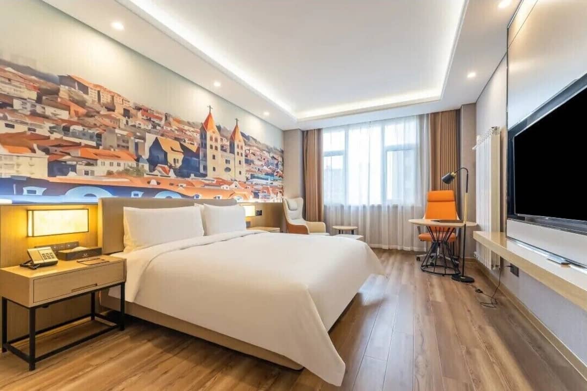 A hotel room with a bed, bedside table, lamp, TV, and a mural of a town on the wall.