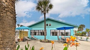A building with a blue exterior, outdoor seating area with chairs and umbrellas, and a palm tree. - Ormond Beach Retreat w/ Deck - Steps to Ocean! (Ormond Beach)