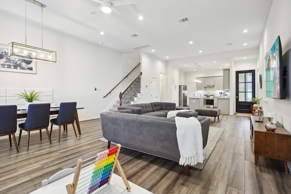A modern living room with a gray sectional sofa, a dining area with chairs, and a kitchen in the background. - Midterm Ready, Large 2King BD Home Near HOU Med CTR, NRG Stadium & HOU Downtown (Houston)