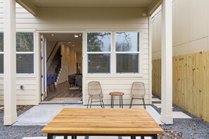 A covered patio with a wooden table and two wicker chairs, a wooden fence, and a view of the house's interior through a glass door. - Midterm Ready, Large 2King BD Home Near HOU Med CTR, NRG Stadium & HOU Downtown (Houston)