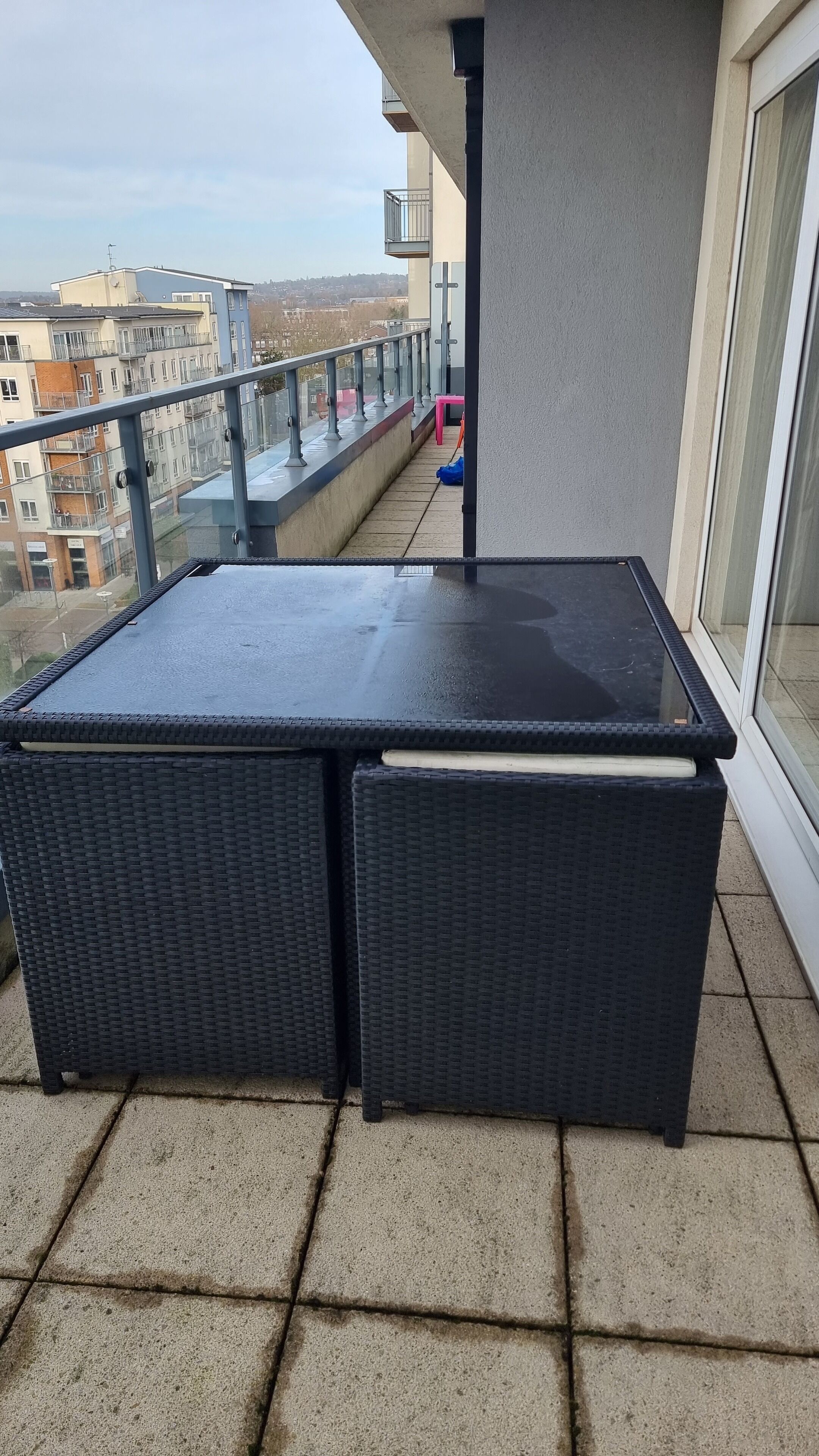 A balcony with a black table and two wicker chairs, a pink chair, and a blue bag on the floor.
