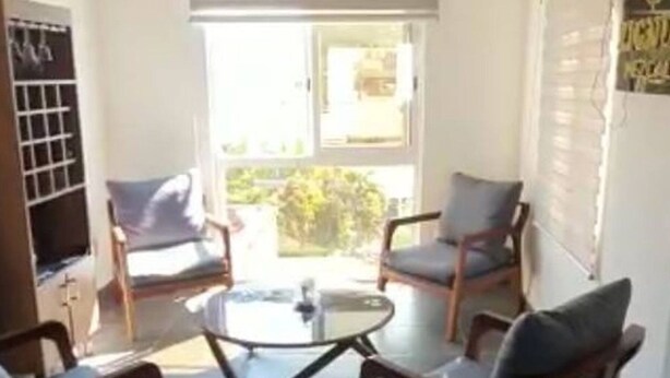 Una habitación con suelo de baldosas, dos sillas de madera con cojines, una mesita y una ventana con persianas.