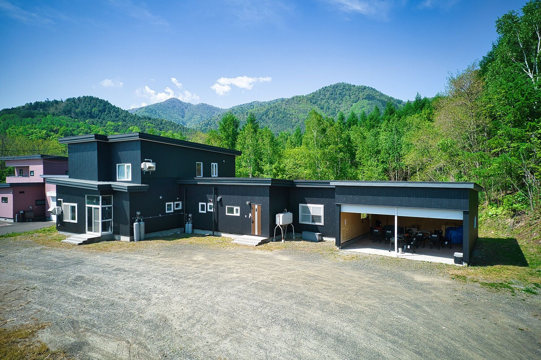 A modern building with a garage and outdoor seating area, surrounded by lush greenery and mountains.