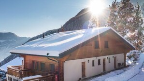 A wooden cabin with a snow-covered roof, a wooden deck, and a mountain backdrop. - Plein Sud 3 by Interhome (Ovronnaz)