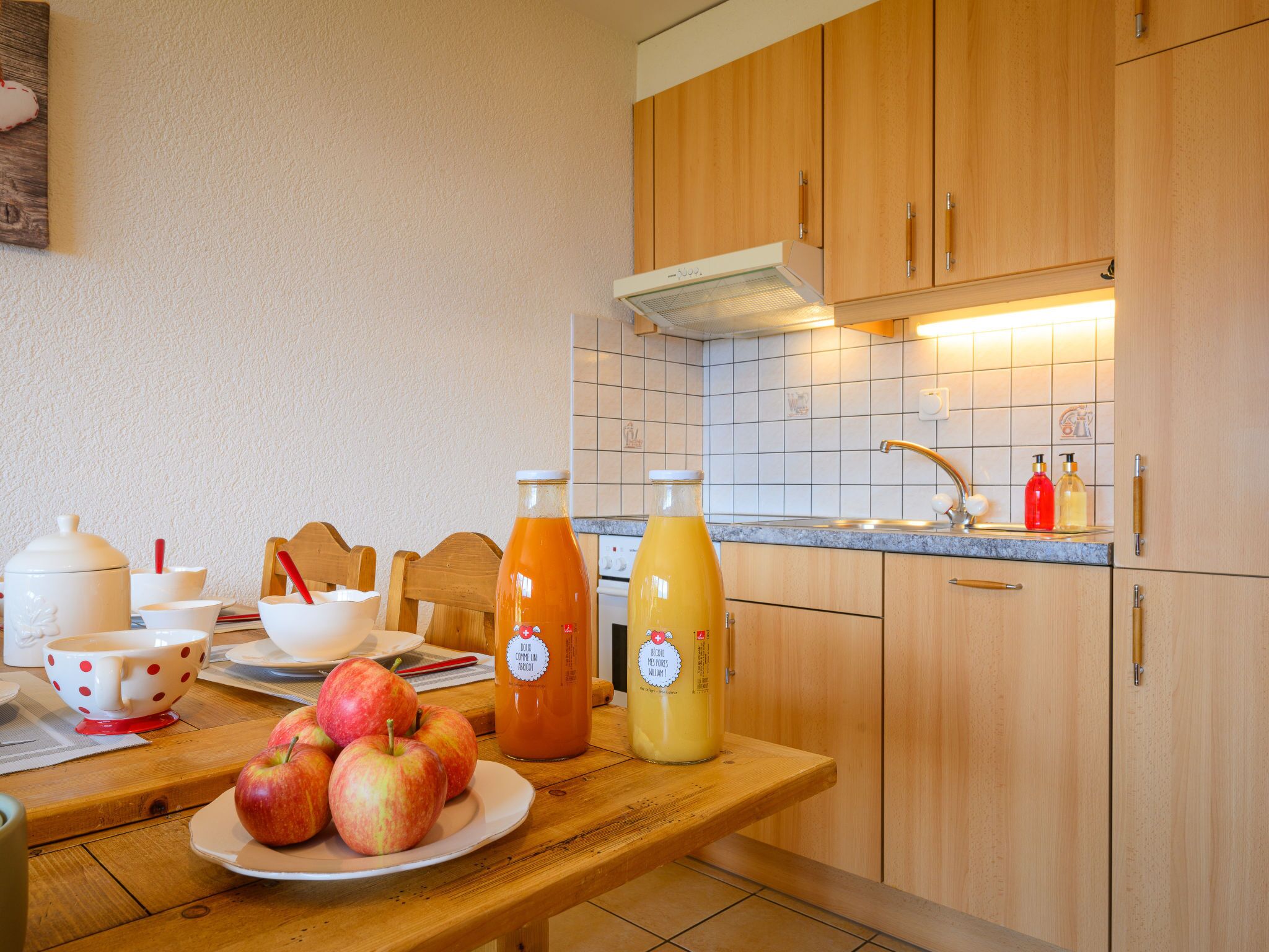 Una cucina con mobili in legno, zoccolo in piastrelle e un tavolo di legno con un piatto di mele e due bottiglie di succo.