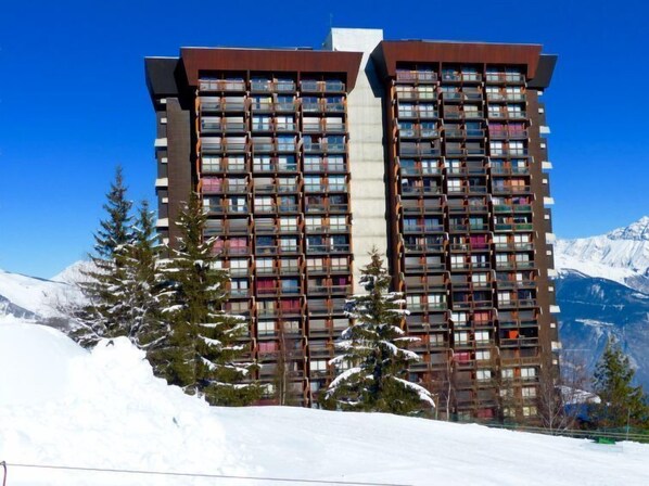 Un hôtel de grande hauteur doté de nombreux balcons, entouré de montagnes enneigées et d’arbres à feuilles persistantes.