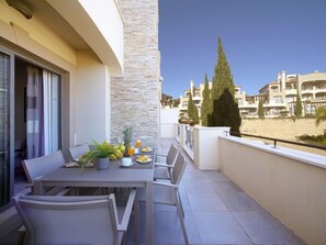 A balcony with a table set for breakfast, including fruits and pastries, overlooking a residential area with buildings and a beach.