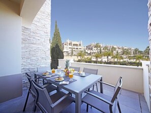 A balcony with a table set for breakfast, including croissants and fruit, overlooking a residential area with palm trees and buildings.