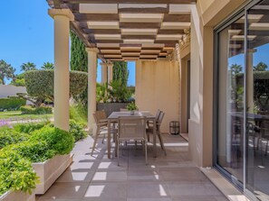 A covered patio area with a table and chairs, surrounded by a garden with various plants and a view of the outdoors.