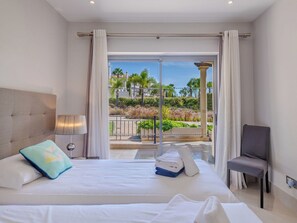 A hotel room with a bed, a chair, a lamp, and a view of a garden and buildings.