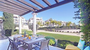 Un patio al aire libre con una mesa lista para comer, rodeado de sillas, y vistas a un césped cuidado y edificios al fondo.