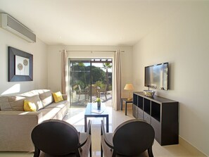 A modern living room with a sofa, chairs, a TV, and a balcony with a view of greenery.
