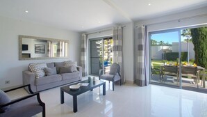 A modern living room with a sofa, a coffee table, and a chair. There is a large window with curtains, and a view of a garden outside. - Pine Hills Pérola by Interhome (Vilamoura)