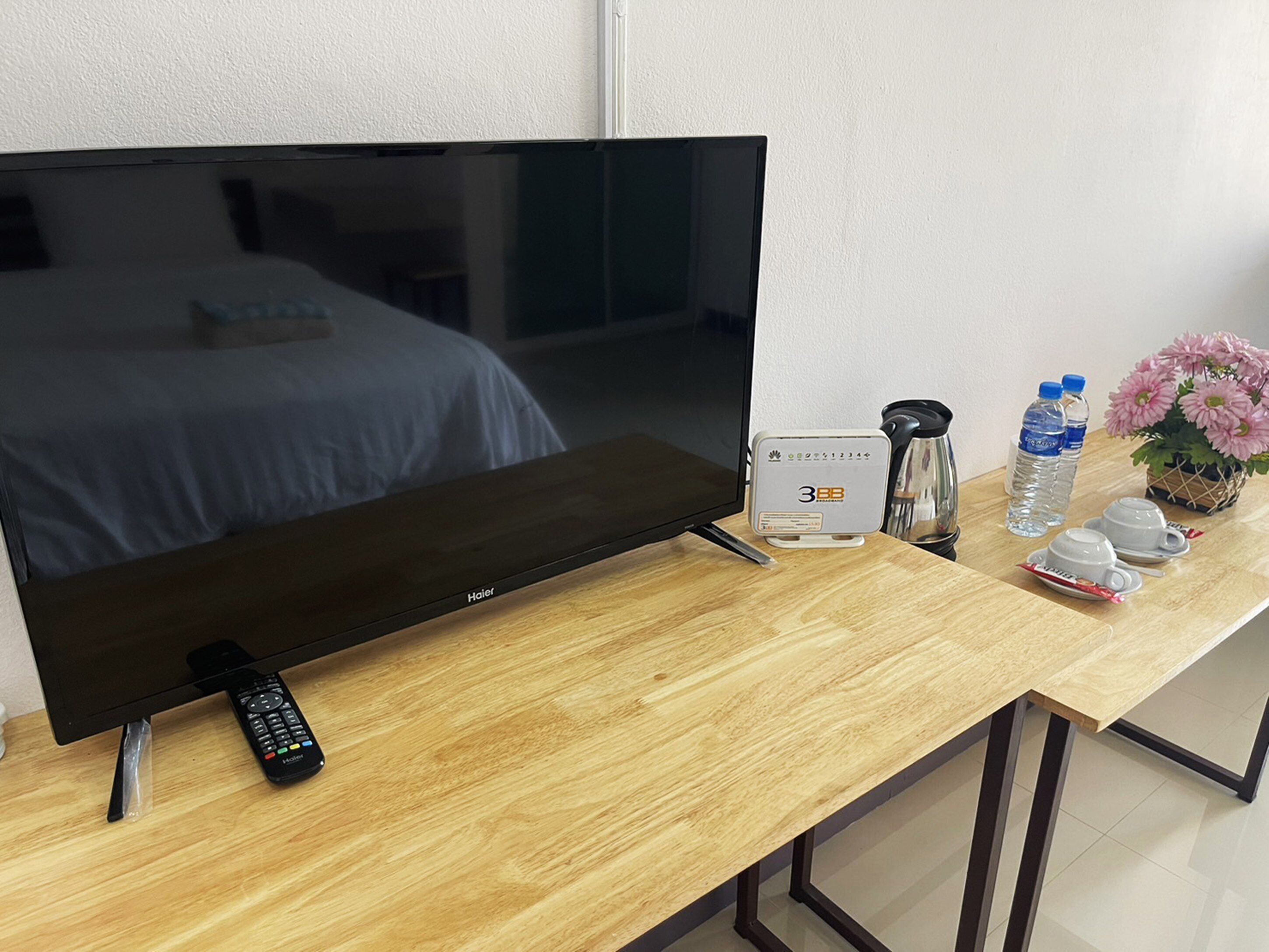 A hotel room with a large flat-screen TV, a bedside table with a coffee maker, water bottles, and a flower arrangement.