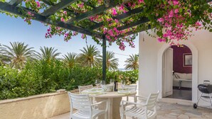 Un patio esterno con un tavolo apparecchiato per mangiare, circondato da sedie bianche, sotto un pergolato con fiori rosa.