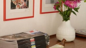 Um quarto com quadros nas paredes, um vaso com flores e uma pilha de revistas sobre a mesa.