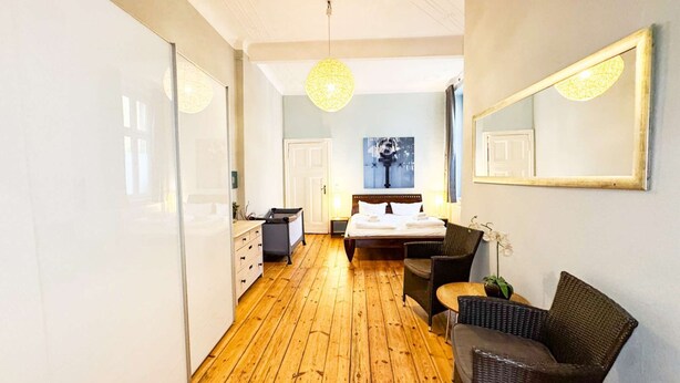 A bedroom with a wooden floor, a bed with white linens, a chair, a small table, and a large mirror on the wall. - primeflats - Apartment Kattegat Berlin-Wedding (Berlin)