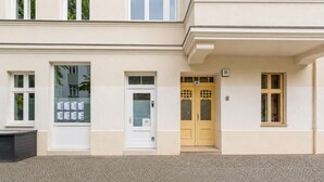 A building with a white facade, a set of double doors with a number plaque, and a window with a bulletin board. - primeflats - Apartment Kattegat Berlin-Wedding (Berlin)