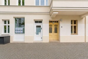 Ein Gebäude mit weißer Fassade, doppelte Türen mit einer Nummertafel und ein Fenster mit einer Aushangtafel.