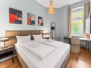 A hotel room with a large bed, two bedside tables, a desk, a chair, a window with curtains, and three abstract paintings on the wall.