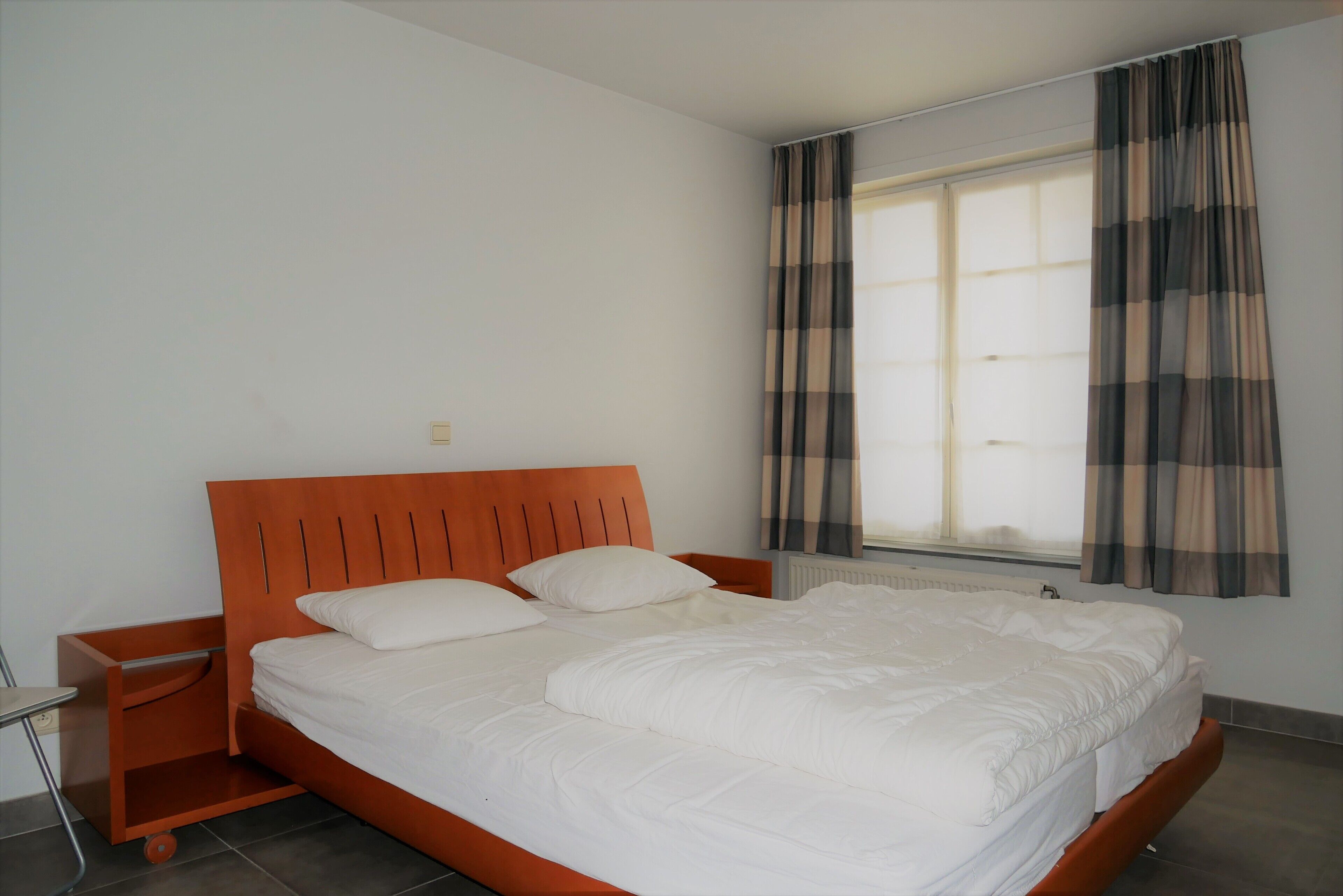 Un dormitorio con cama de madera, ropa de cama blanca, una mesita de noche y una ventana con cortinas.