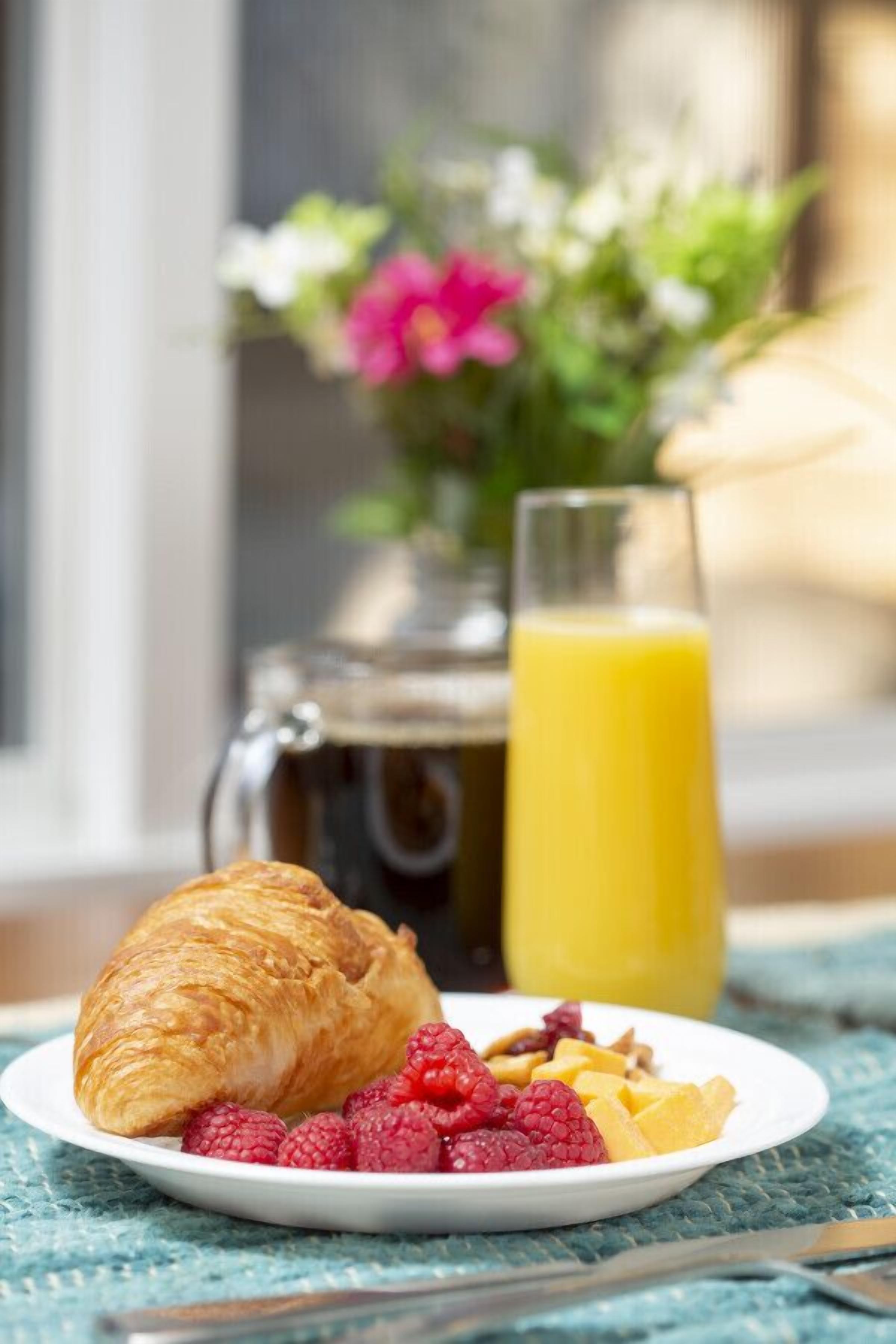 A breakfast setting with a croissant, raspberries, and orange segments on a plate, a glass of orange juice, and a coffee pot in the background.