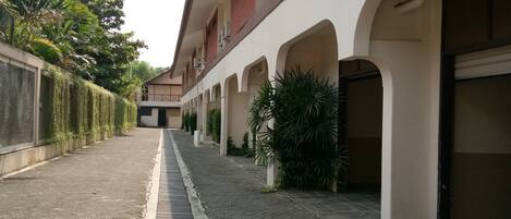 A covered walkway with a brick floor, bordered by a low wall on one side and a building with arched windows and air conditioning units on the other.