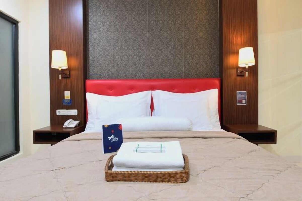 A hotel room with a red upholstered headboard, white bedding, and two bedside lamps. A wicker basket with folded towels is on the bed.