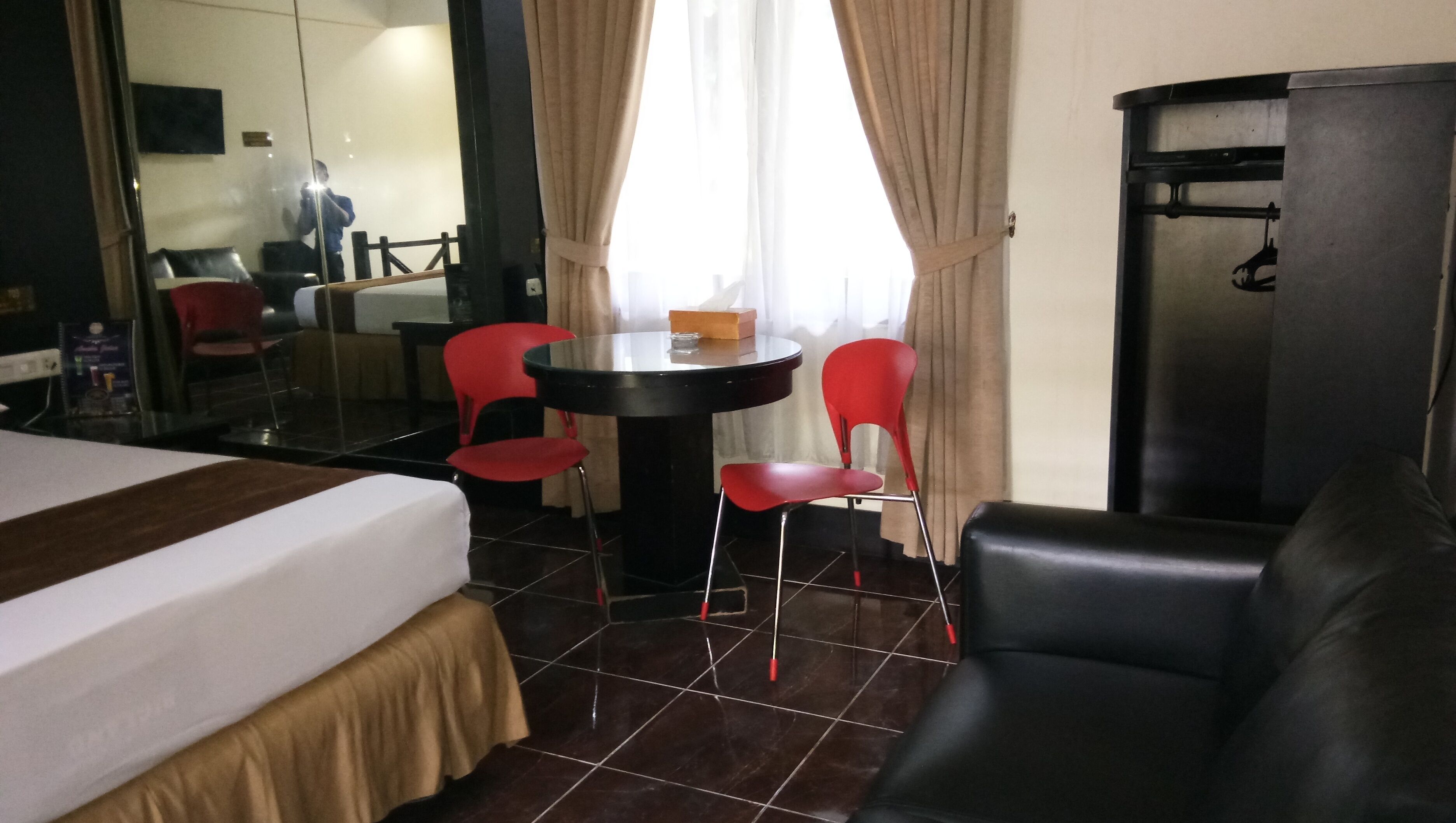 A hotel room with a bed, a round table with two red chairs, a black leather sofa, and a minibar.