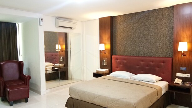Une chambre d’hôtel avec un grand lit, un fauteuil rouge recouvert de tissu, une coiffeuse avec un miroir et deux lampes de chevet.