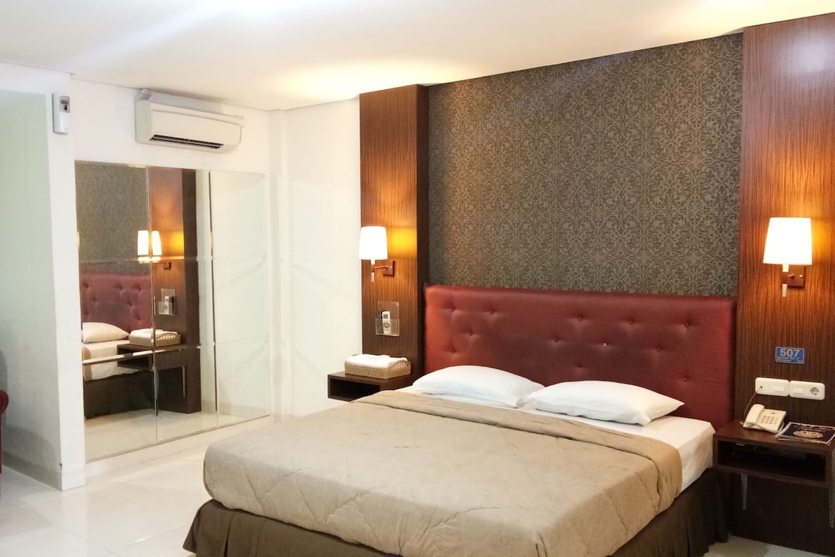 Une chambre d’hôtel avec un grand lit, un fauteuil rouge recouvert de tissu, une coiffeuse avec un miroir et deux lampes de chevet.