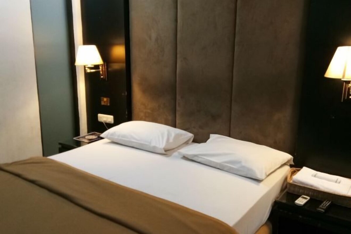 A hotel room with a bed, two bedside lamps, a nightstand with a phone, and a brown throw blanket.