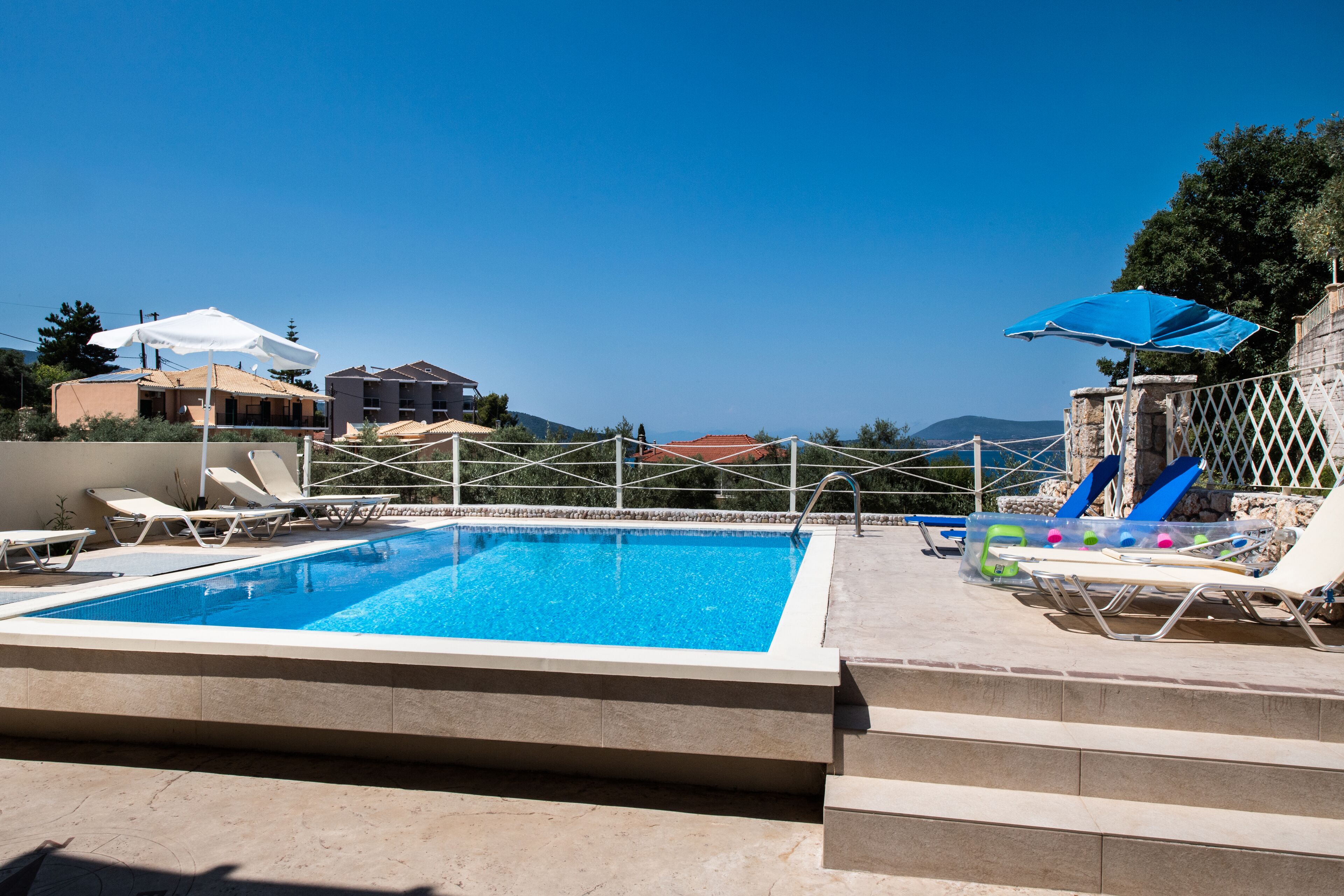 A swimming pool with lounge chairs and umbrellas, surrounded by buildings and a clear sky.