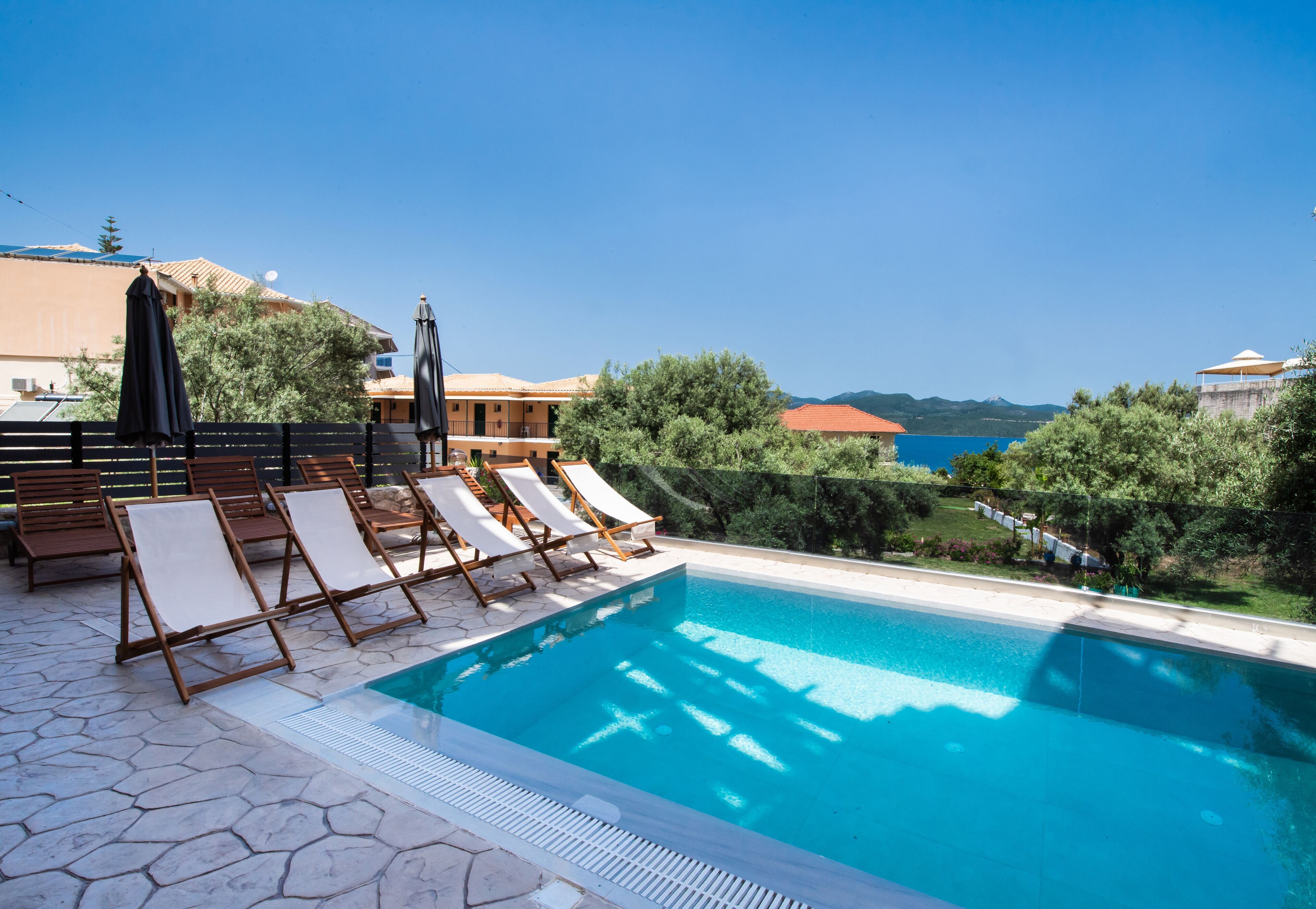 A pool area with lounge chairs and umbrellas, surrounded by greenery and a view of the sea.