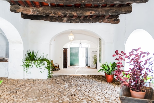 A courtyard with a cobblestone floor, white walls, and wooden beams. There are potted plants and a doorway leading to another room. - Eighteen21 Houses - Casa dos Condes (Sousel)
