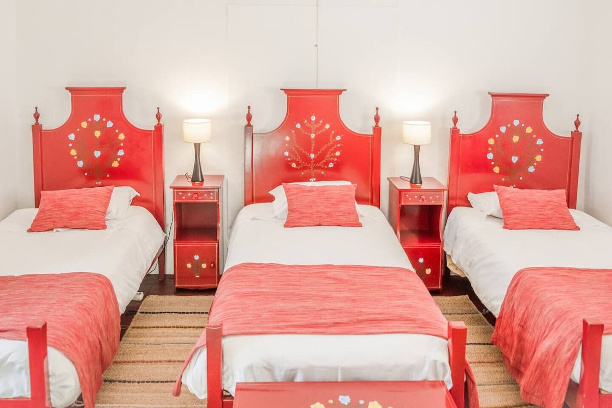 A hotel room with three beds, each with a red headboard and white bedding. There are bedside tables with lamps and decorative items.
