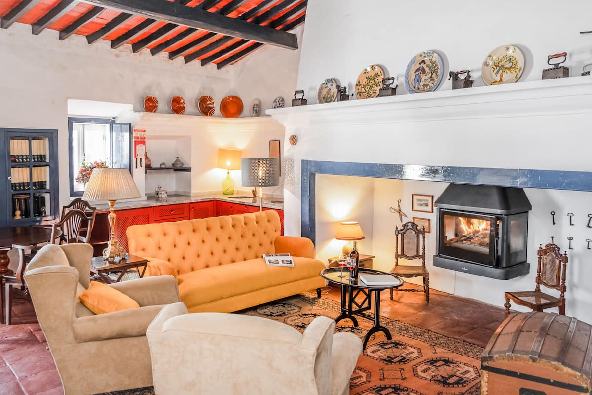 A cozy living room with a fireplace, a sofa, and a coffee table. There is a bookshelf and a dining area in the background.
