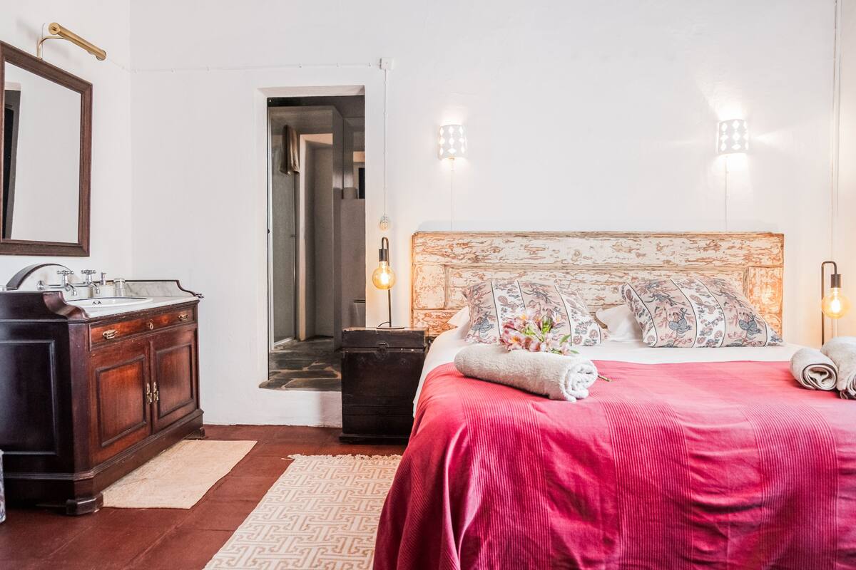 A bedroom with a wooden headboard, a bed with a red bedspread, a nightstand with a lamp, a dresser, and a mirror.