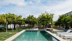 A rectangular swimming pool with clear water, surrounded by a grassy area with trees and a pergola structure. There are lounge chairs along the poolside. - Eighteen21 Houses - Casa dos Condes (Sousel)