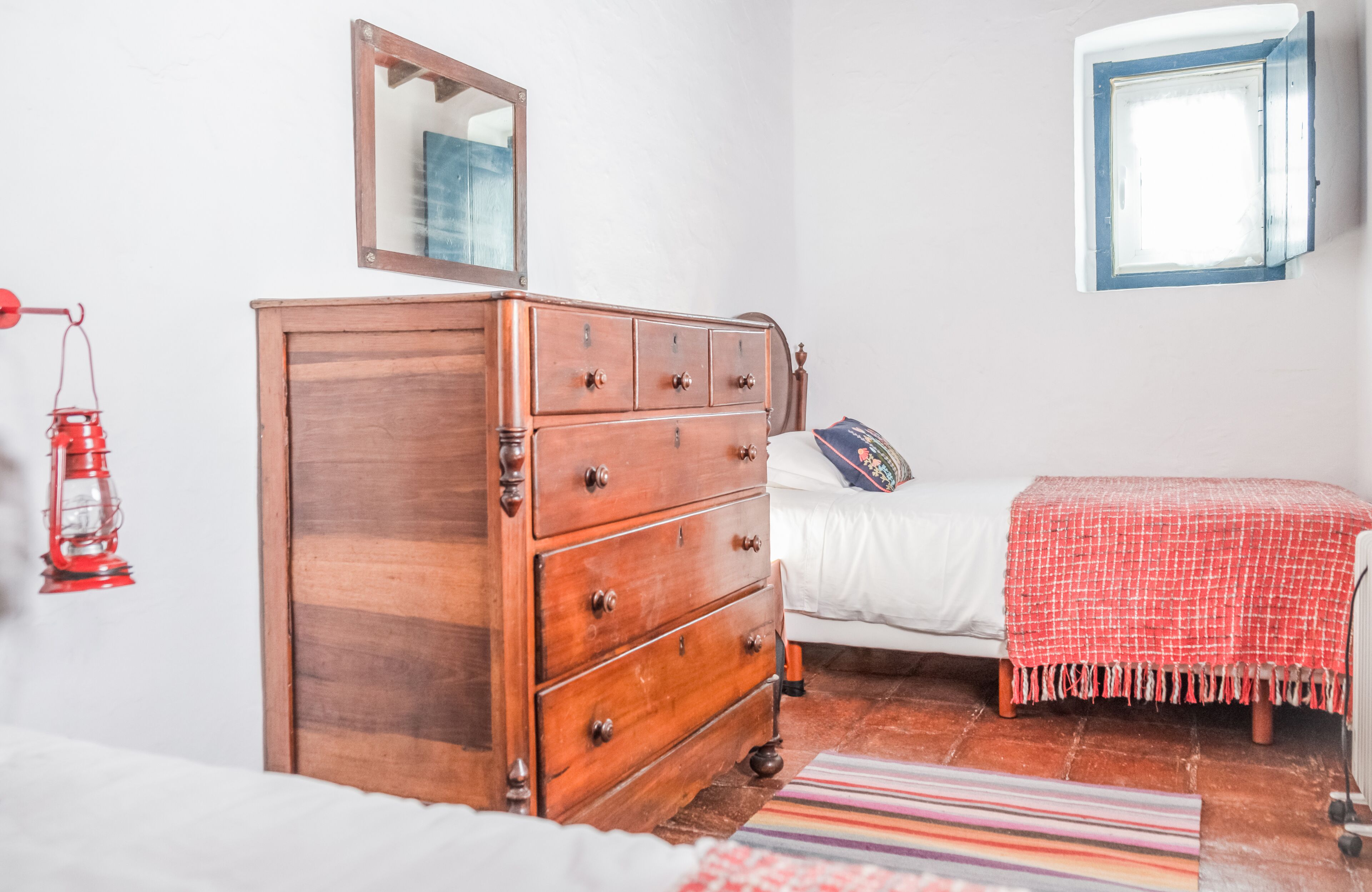 A bedroom with a wooden dresser, a bed with a white comforter, a red lantern, and a window with a blue frame.