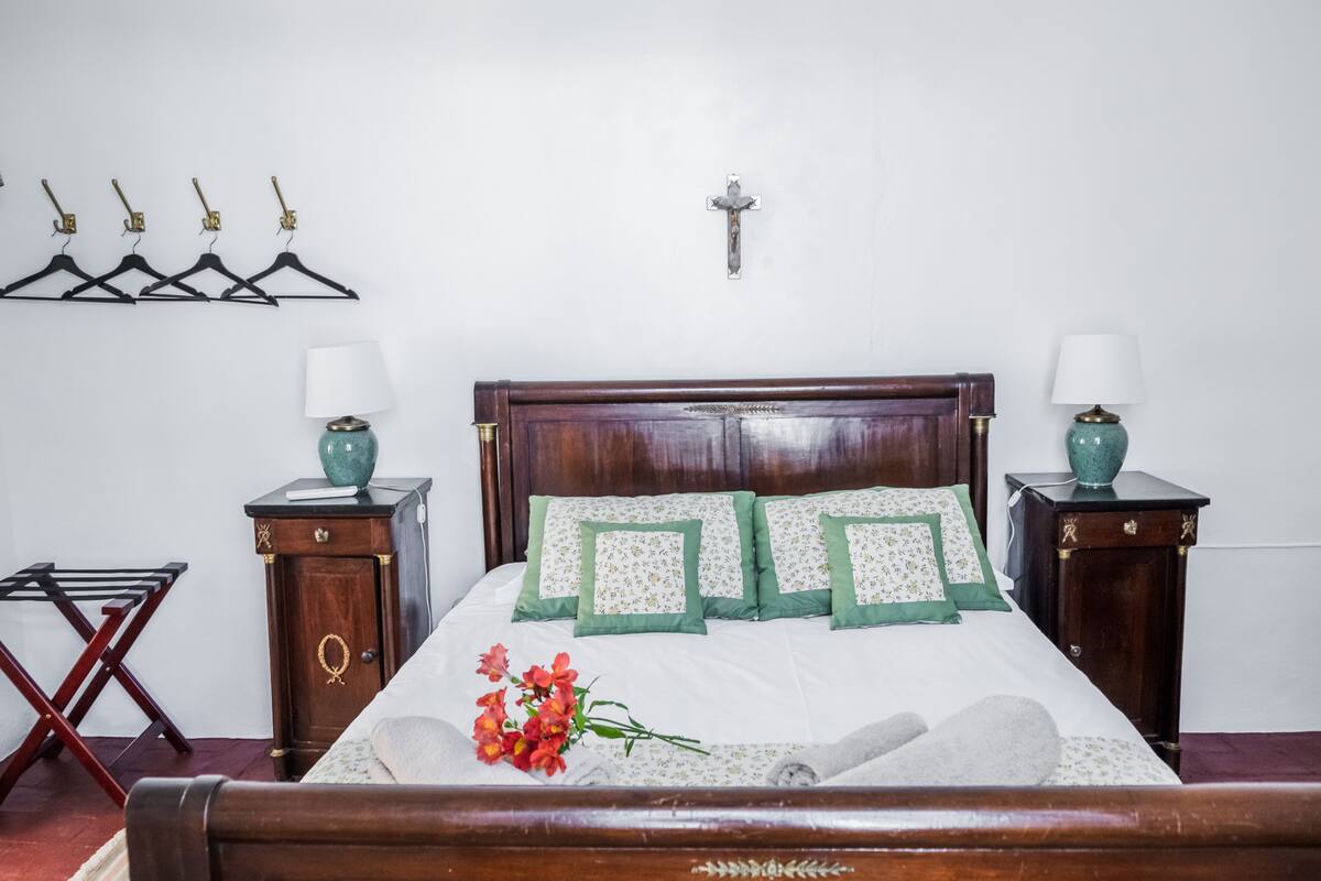 A neatly made bed with green and white pillows, a wooden headboard, and a crucifix on the wall. There are two bedside tables with lamps and a drying rack.