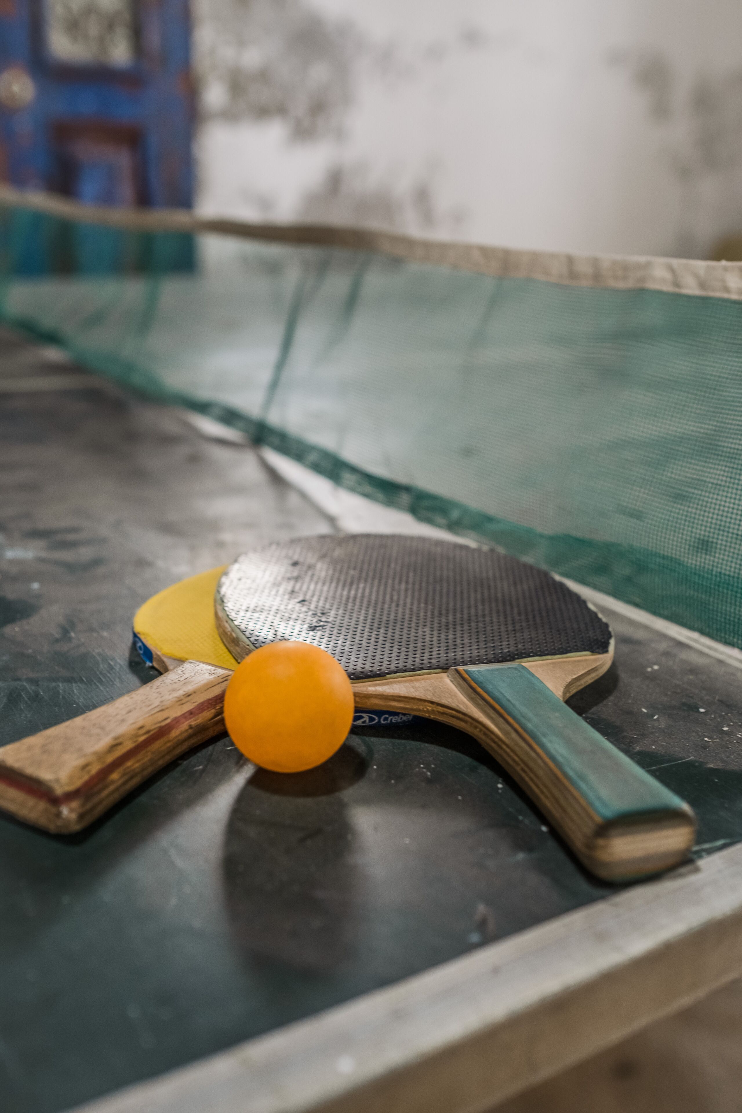Une table de ping-pong avec un filet, une raquette jaune, une raquette en bois et une balle jaune.