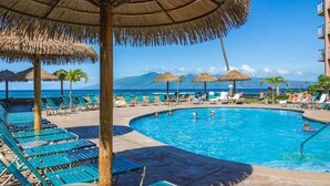 A pool area with lounge chairs and thatched umbrellas, surrounded by a clear blue sky and a mountain view.