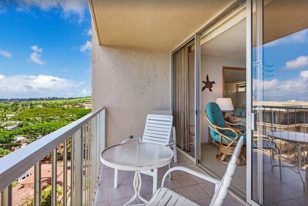 Un balcón con mesa y sillas, vistas a vegetación y un vistazo a una habitación con lámpara y sillón.
