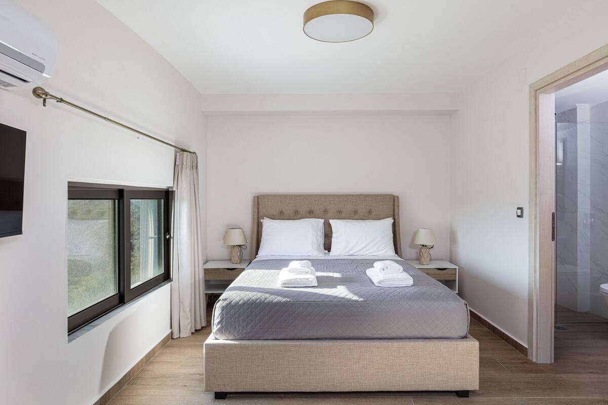A modern bedroom with a large bed, bedside tables, a window, and a bathroom visible through an open door.