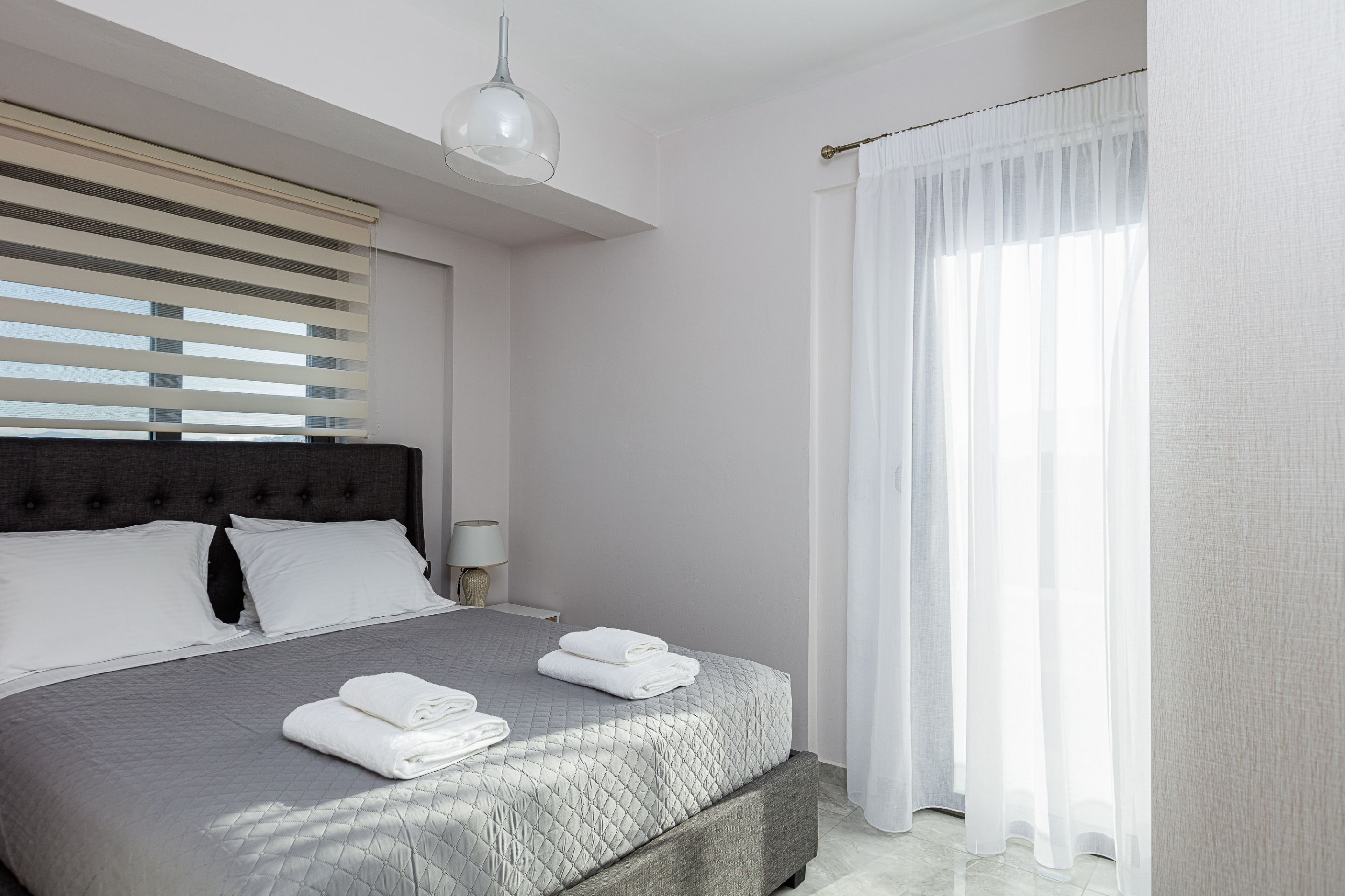 A neatly made bed with folded towels, a modern headboard, and sheer curtains on a window.
