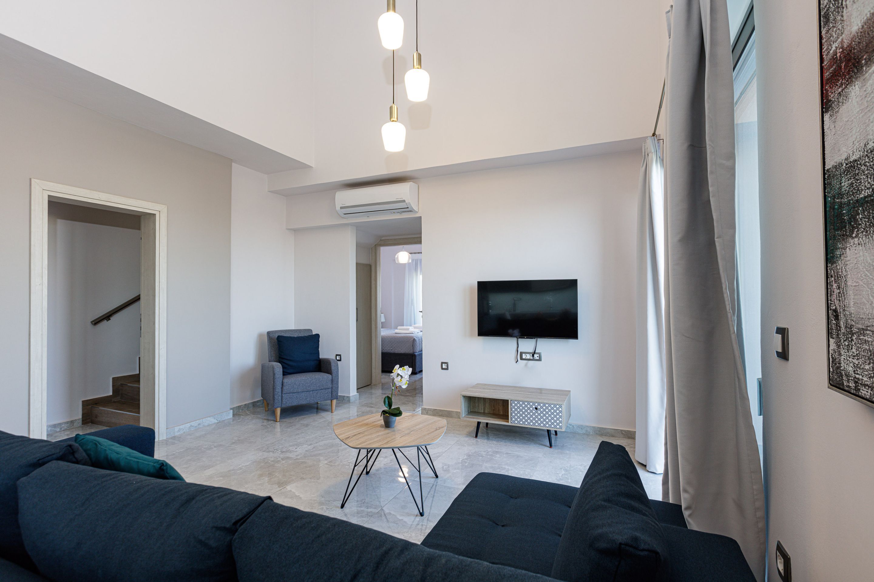 Een moderne woonkamer met een donkere bank, een salontafel, een televisie en een trap naar een andere kamer.